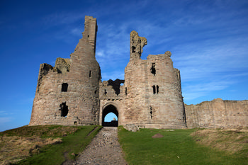 Dunstanburgh castle gate This architecture photograph shows the gatehouse of Dunstanburgh Castle, a historic coastal fortress located in Northumberland, England, United Kingdom. Captured in the early afternoon during winter, the image highlights the weathered stone structure set against a blue sky, with the low winter sun casting minimal shadows across the grassy approach. Dunstanburgh Castle stands on the Northumberland coast and is cared for by English Heritage, making it a notable landmark in the region. The ruins, consisting of the twin towers and arched entrance, illustrate the defensive design typical of medieval castles in England.